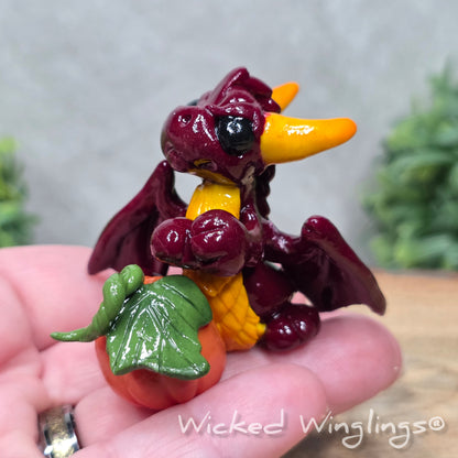 A hand holding a baby clay dragon sculpture with a pumpkin against a wood background with greenery and text Wicked Winglings