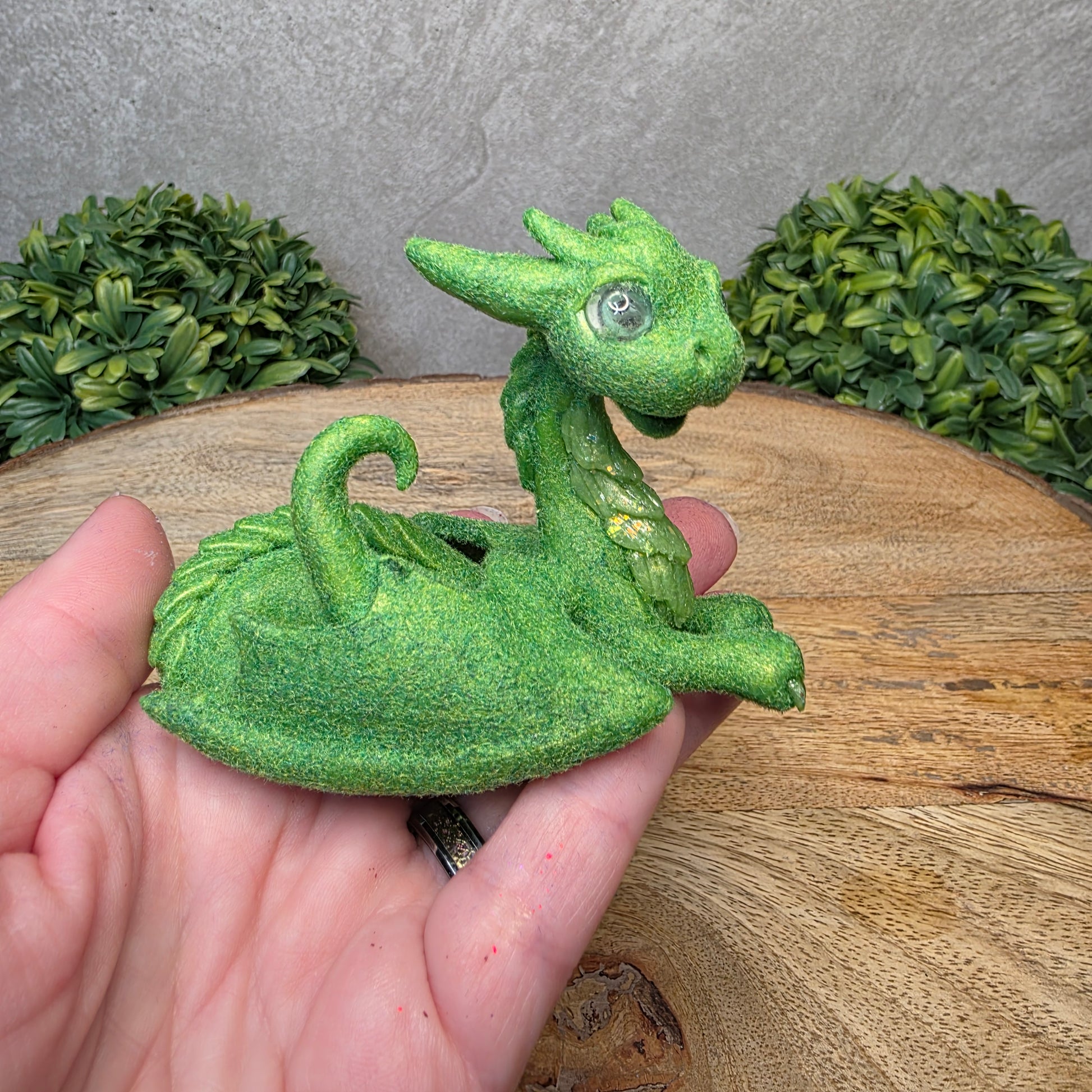 A hand holding a flocked clay dragon sculpture on a wood surface with greenery.
