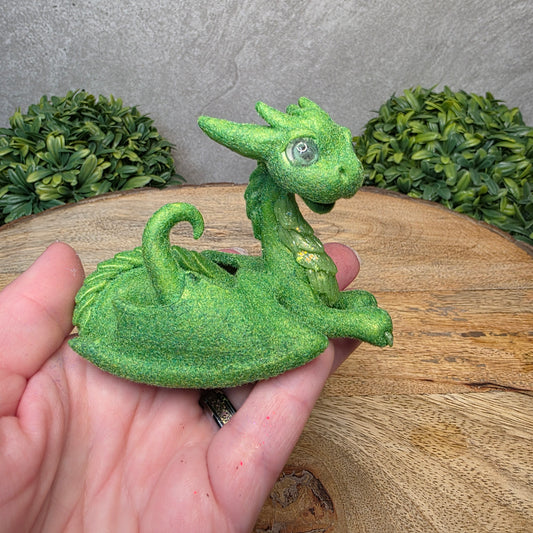 A hand holding a flocked clay dragon sculpture on a wood surface with greenery.