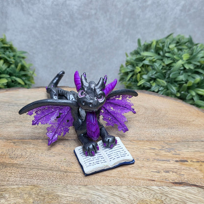 A dragon reading a book against a wood surface with greenery