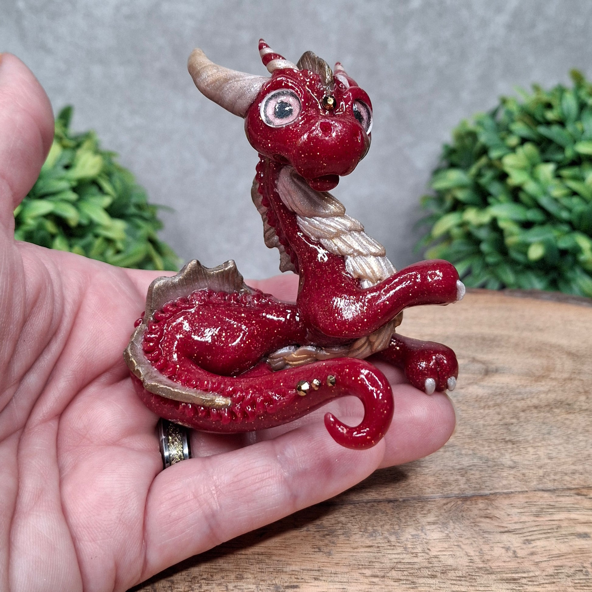 A hand holding a clay dragon against a wood background with greenery