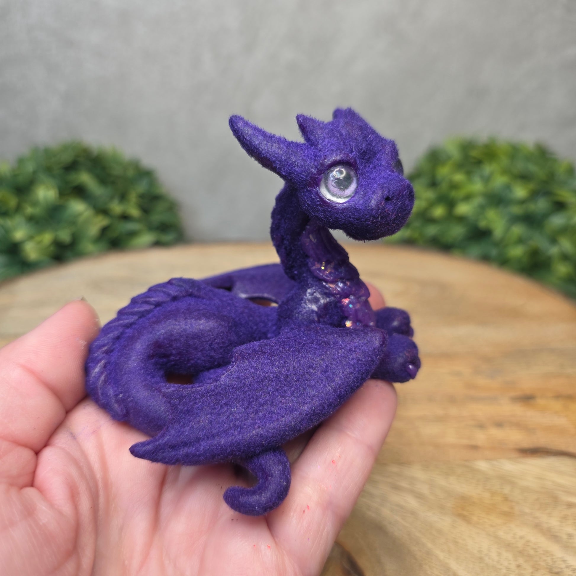 A hand holding a flocked clay dragon sculpture on a wood surface with greenery.