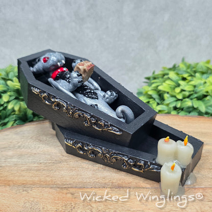 A clay dragon in a black wood coffin with candles and a skull on a wood surface with greenery