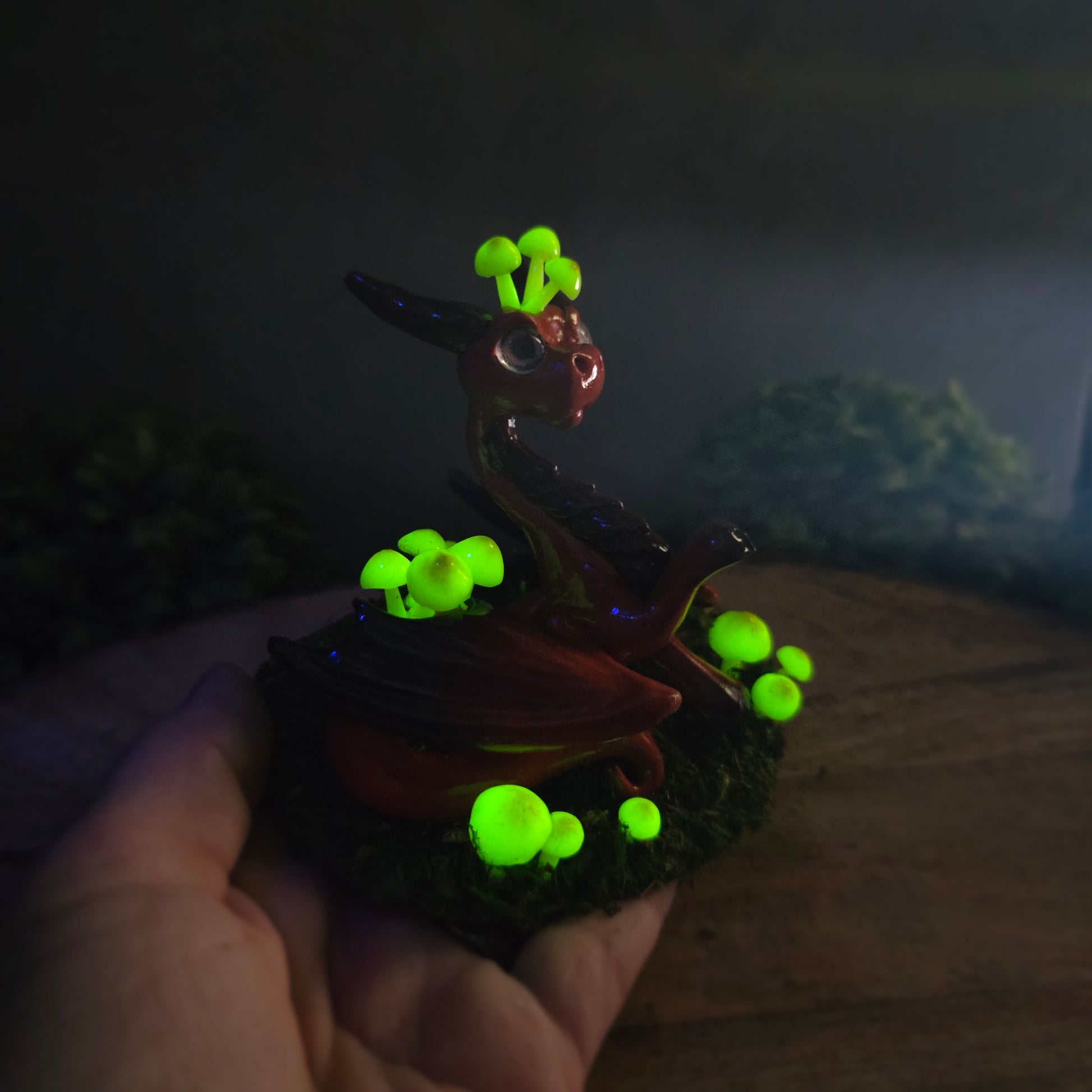 Hand holding a clay dragon figure in dark with glowing mushrooms