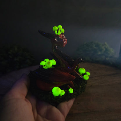 Hand holding a clay dragon figure in dark with glowing mushrooms