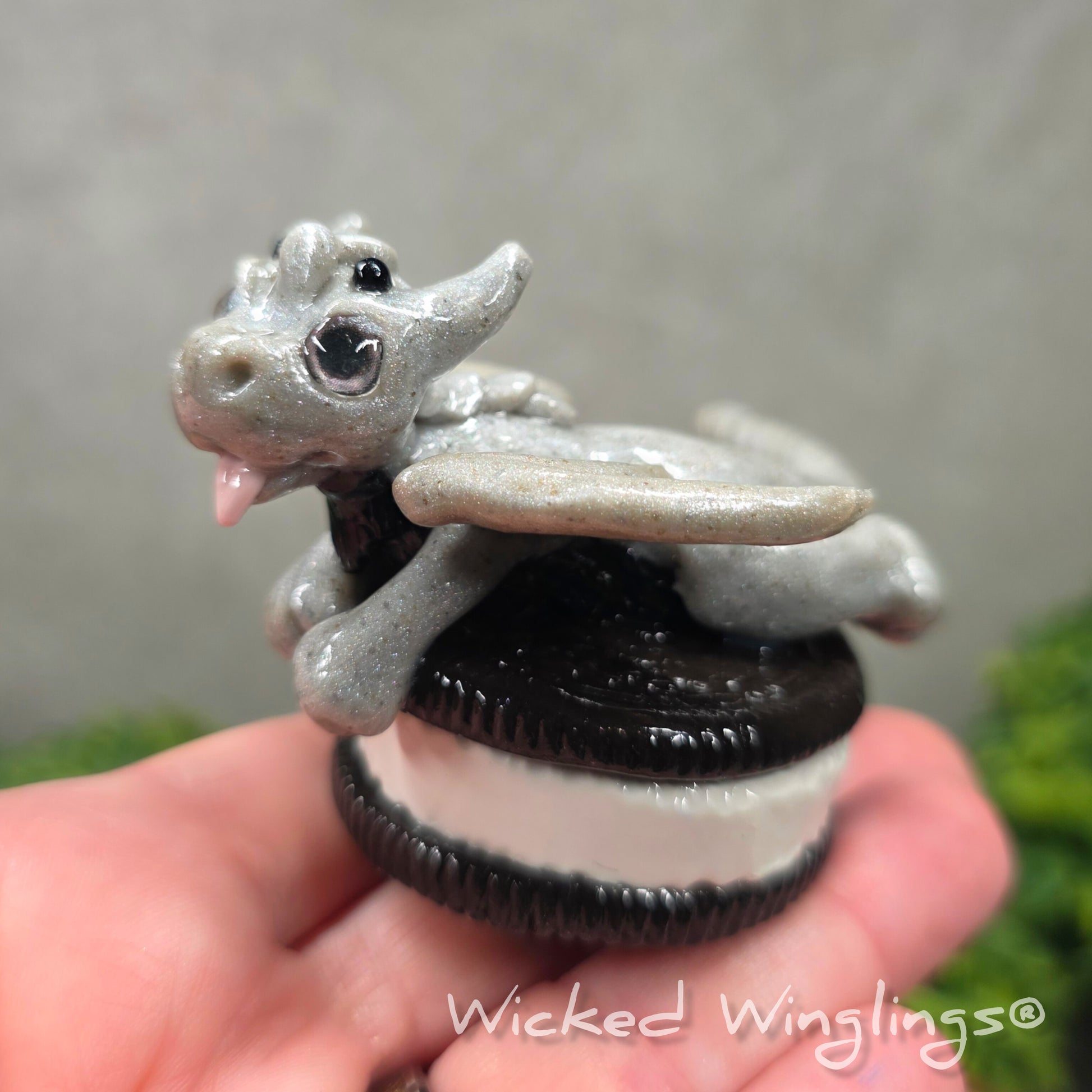 A hand holding a baby clay dragon with a cookie with a Wicked Winglings® watermark.