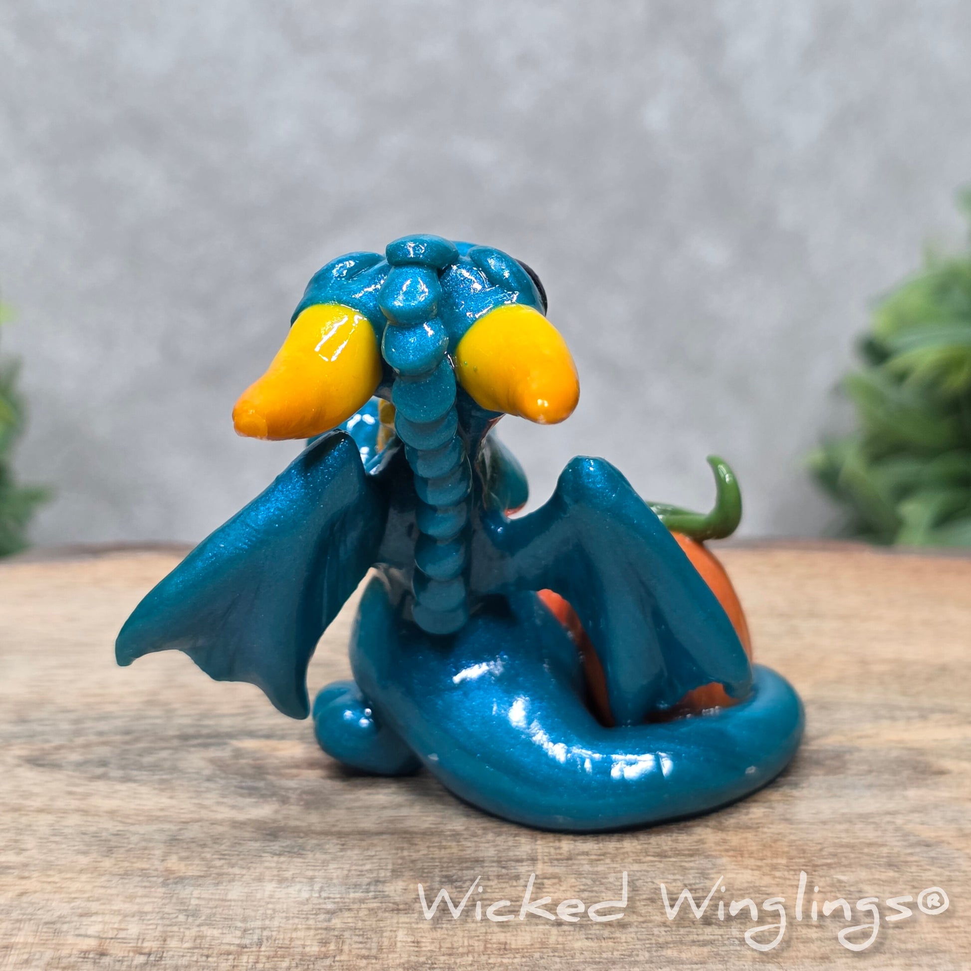 A baby clay dragon sculpture with pumpkins against a wood background with greenery and text Wicked Winglings