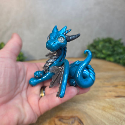 A hand holding a blue and graphite clay dragon against a blurred wood surface