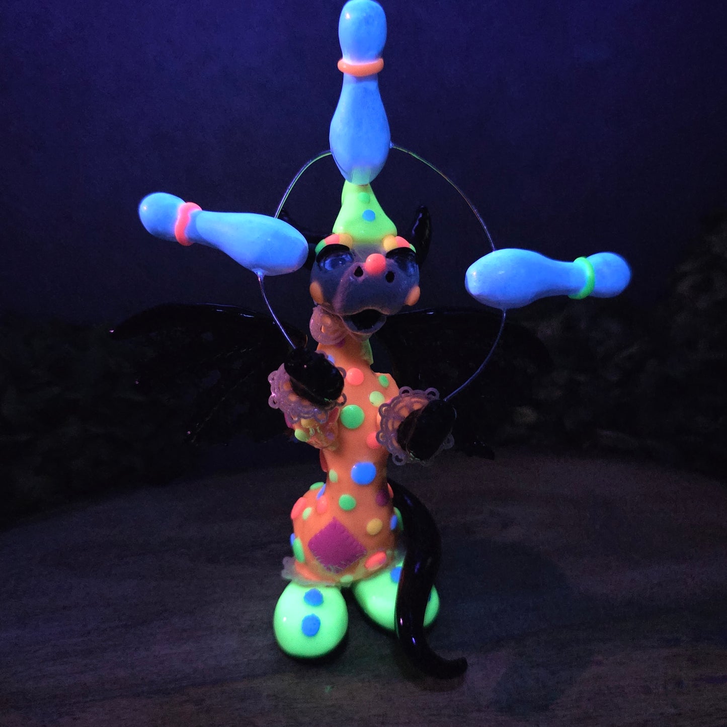 A blacklight reactive clown dragon juggling on a wood surface with greenery