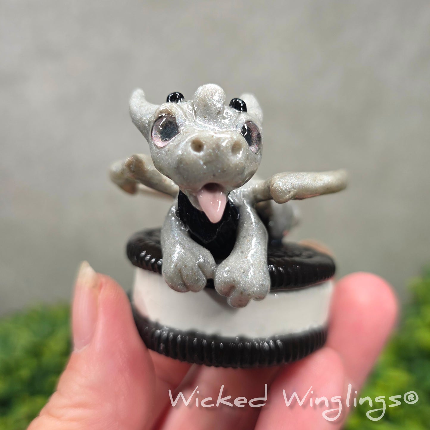 A hand holding a baby clay dragon with a cookie with a Wicked Winglings® watermark.