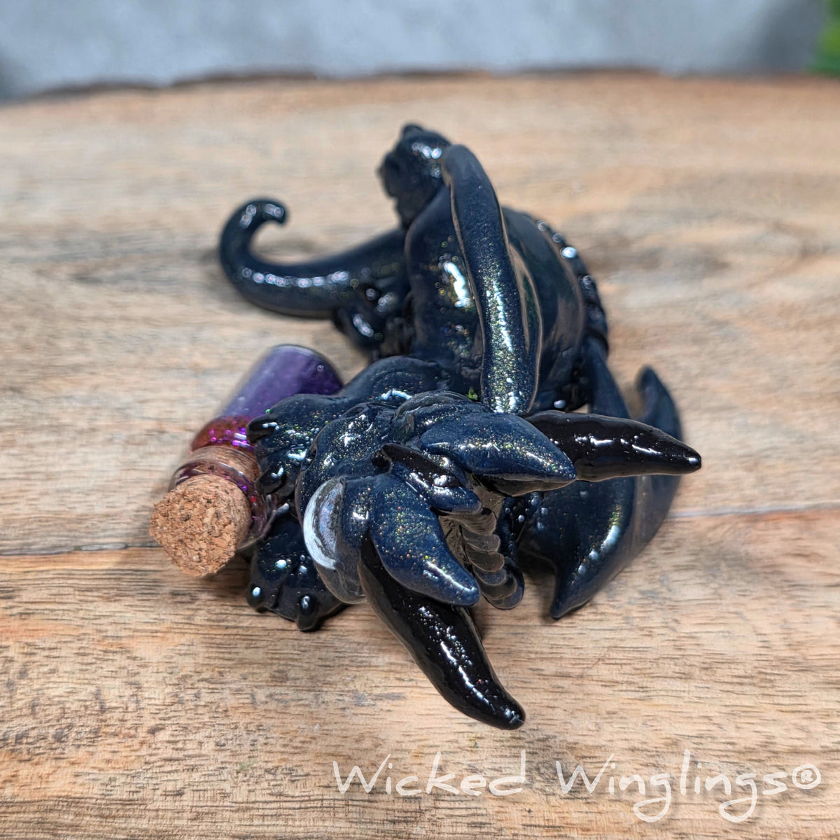 A clay dragon with a potion bottle on a wood surface. Text reads Wicked Winglings.