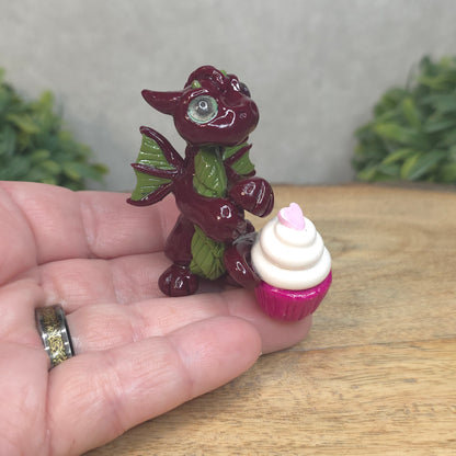A hand holding a baby polymer clay dragon with a cupcake  against a blurred background