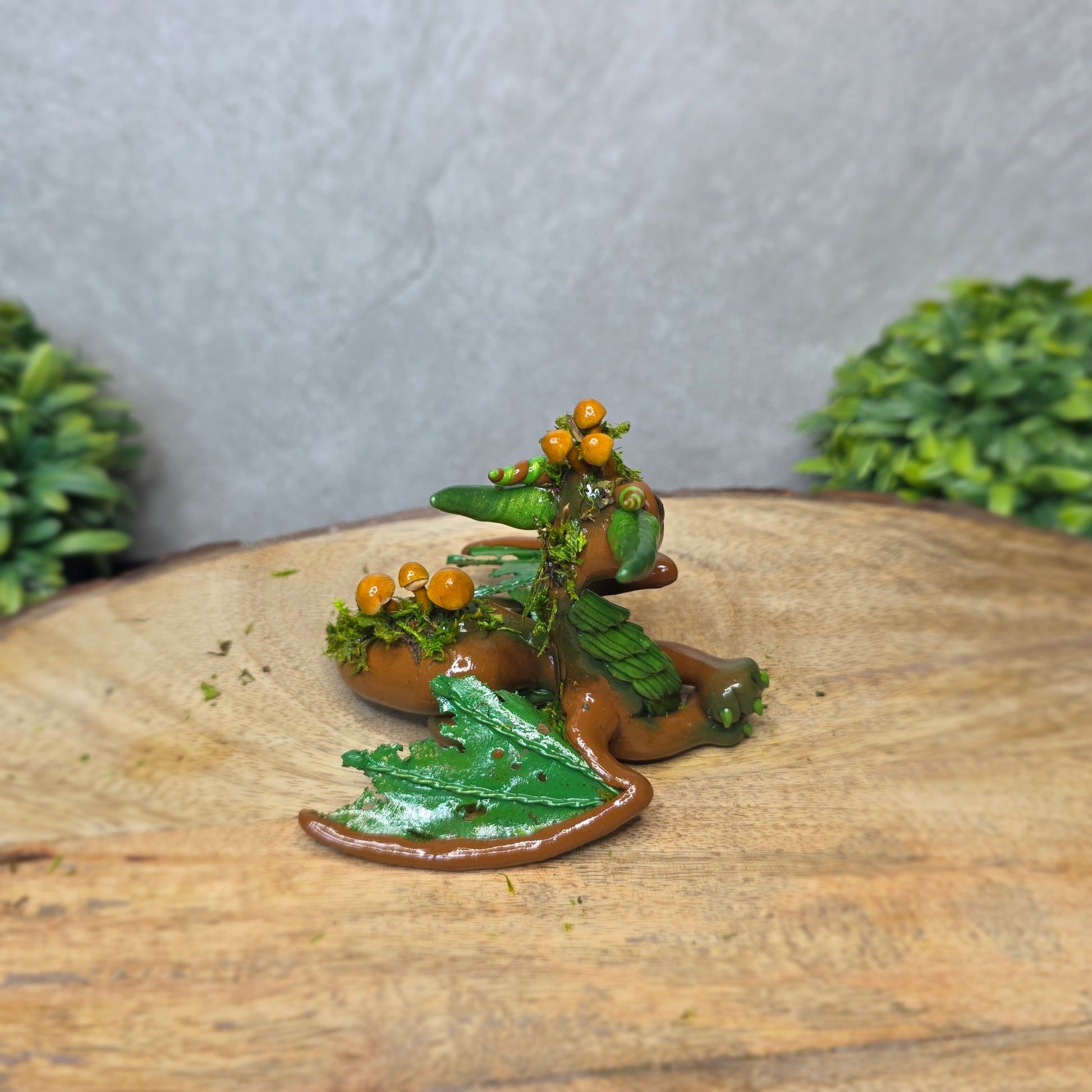 A clay dragon accented with moss and mushrooms against a wood surface with greenery.