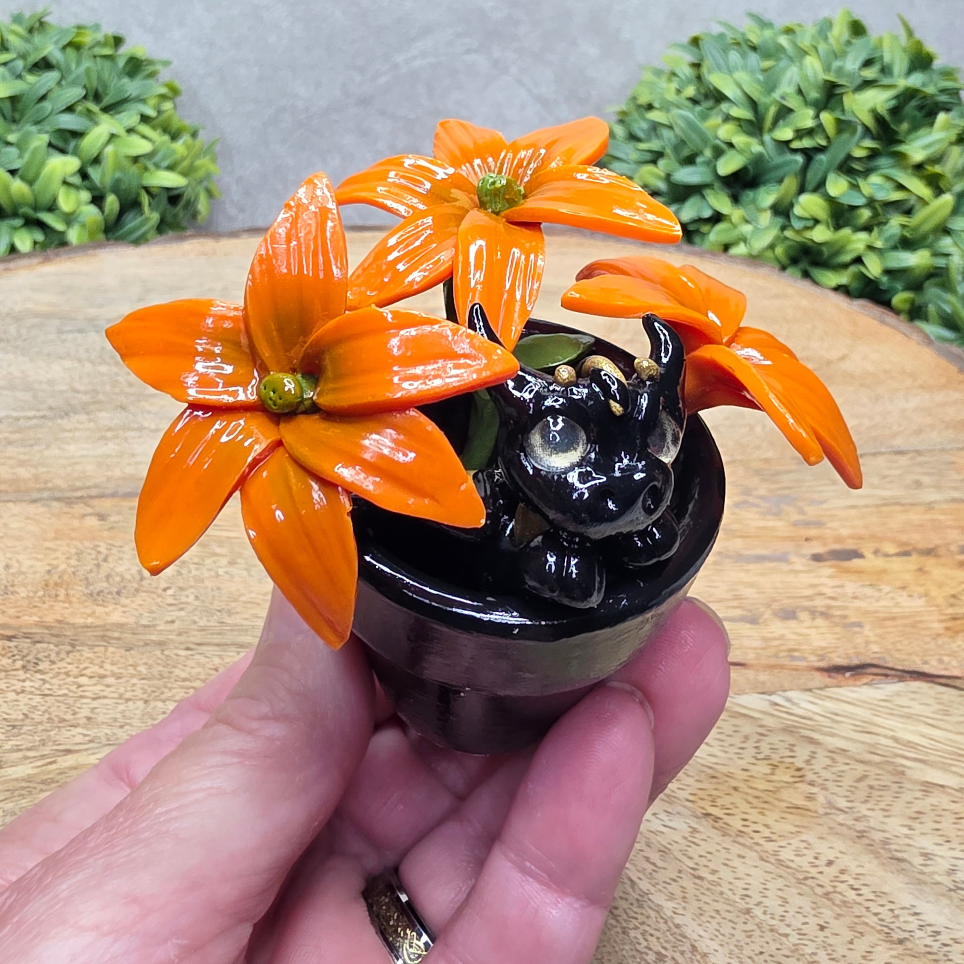 Hand holding a clay baby dragon in a pot with lilies against a wood surface with greenery.