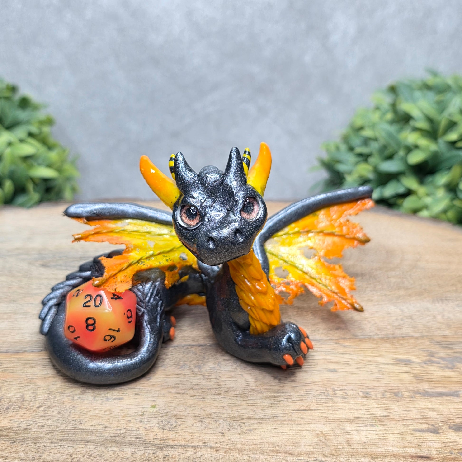 A clay dragon with a D20 dice against a wood surface with greenery.