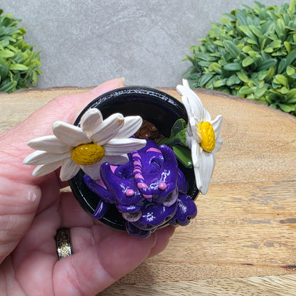 Top view of a clay baby dragon in a pot with daisies against a wood surface with greenery.