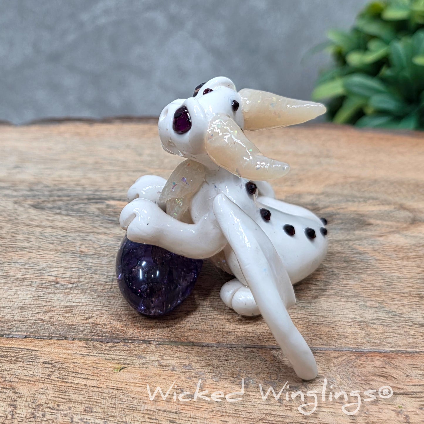 A baby clay dragon sculpture with a crackled quartz against a wood background with greenery and text Wicked Winglings