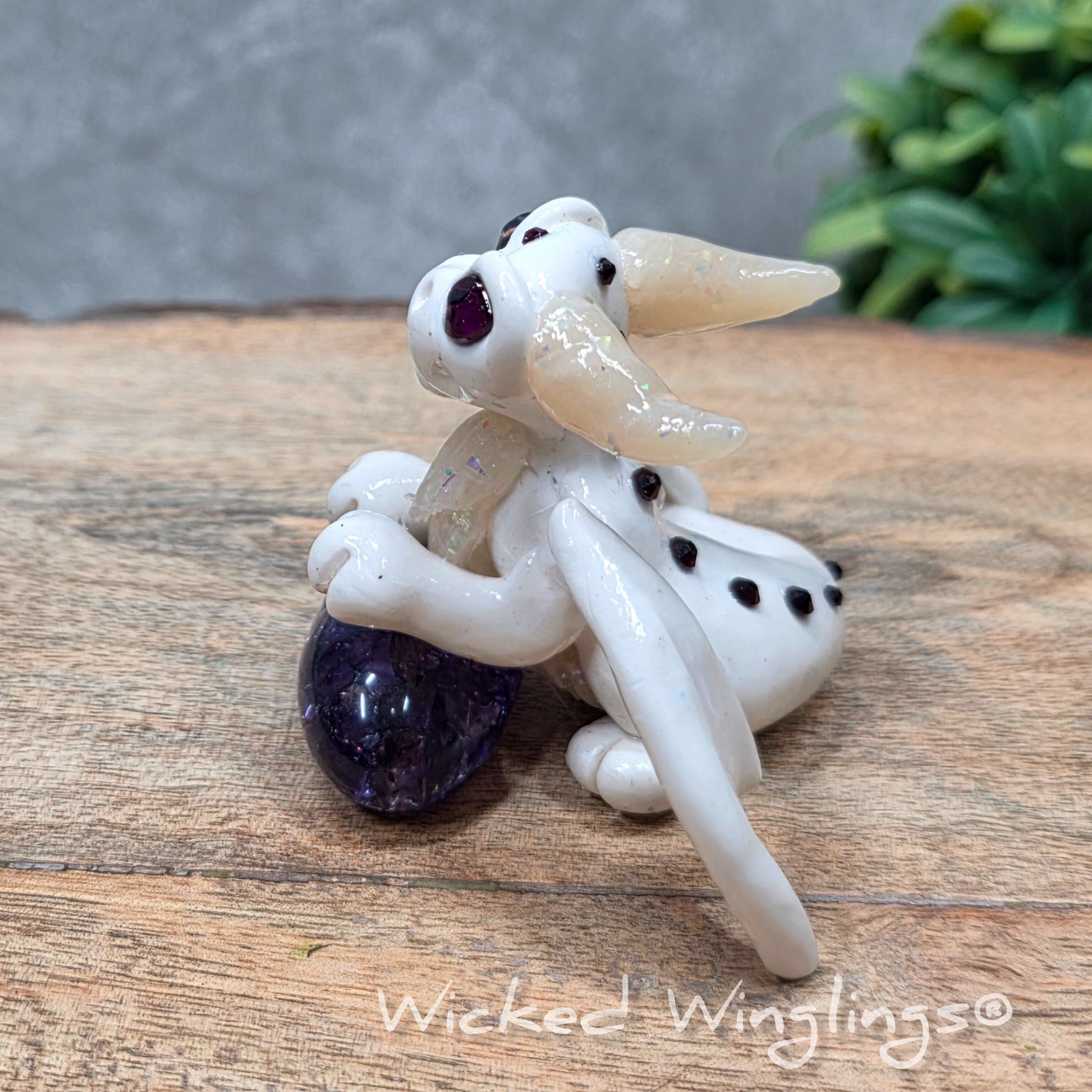 A baby clay dragon sculpture with a crackled quartz against a wood background with greenery and text Wicked Winglings