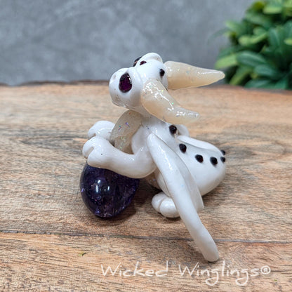 A baby clay dragon sculpture with a crackled quartz against a wood background with greenery and text Wicked Winglings