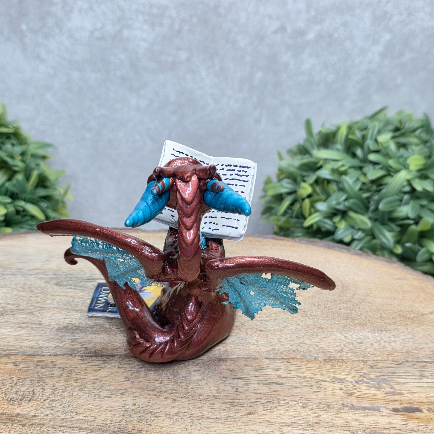 A dragon reading a book against a wood surface with greenery