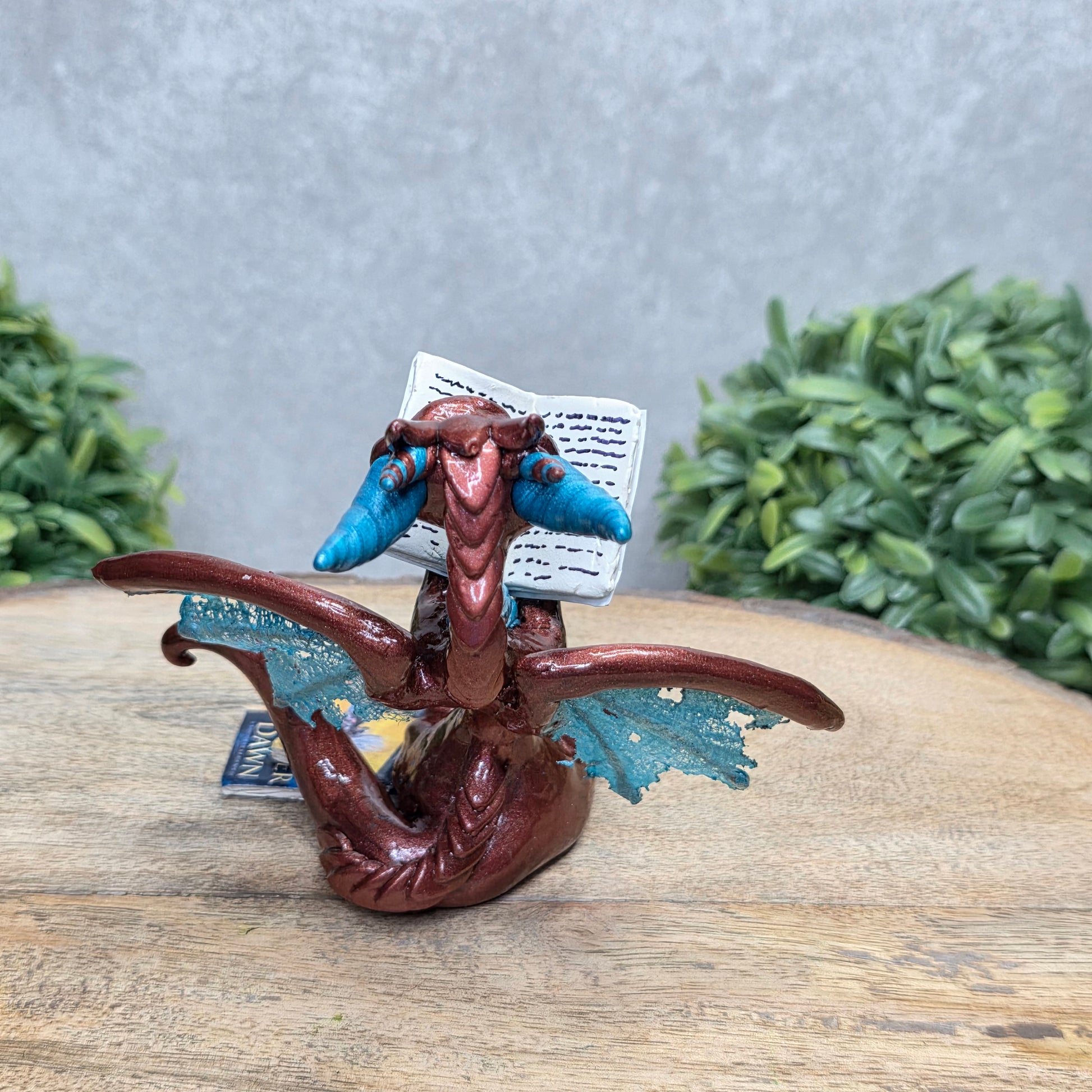 A dragon reading a book against a wood surface with greenery