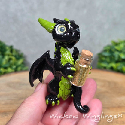 A hand holding a clay dragon with a potion bottle on a wood surface. Text reads Wicked Winglings.