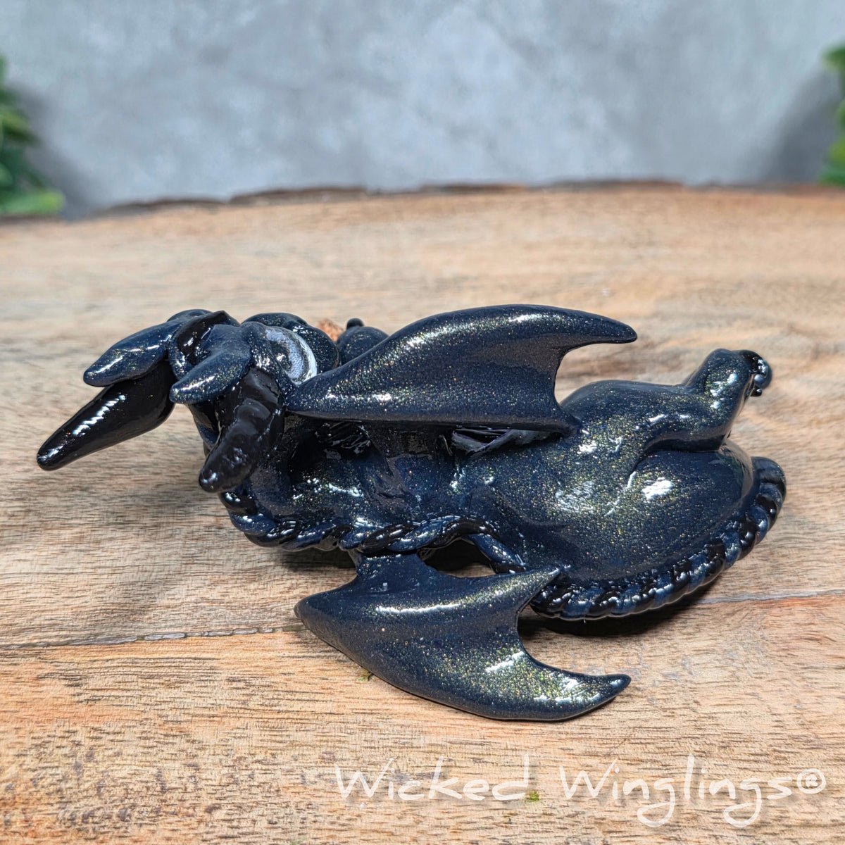 A clay dragon with a potion bottle on a wood surface. Text reads Wicked Winglings.