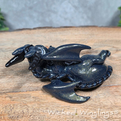 A clay dragon with a potion bottle on a wood surface. Text reads Wicked Winglings.