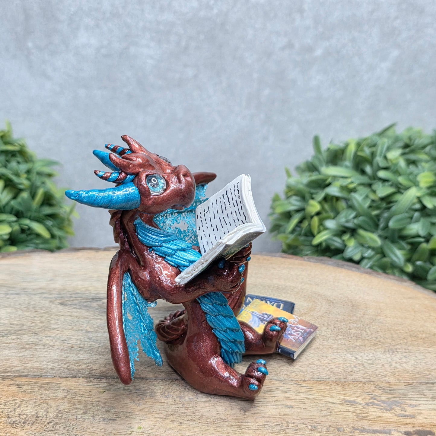 A dragon reading a book against a wood surface with greenery