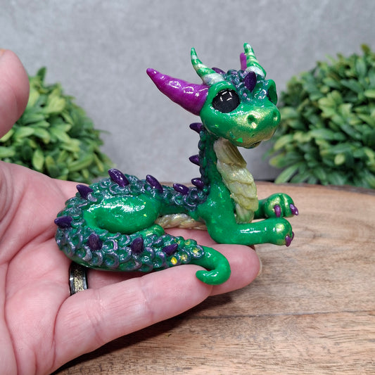 A hand holding a clay dragon against a wood background with greenery