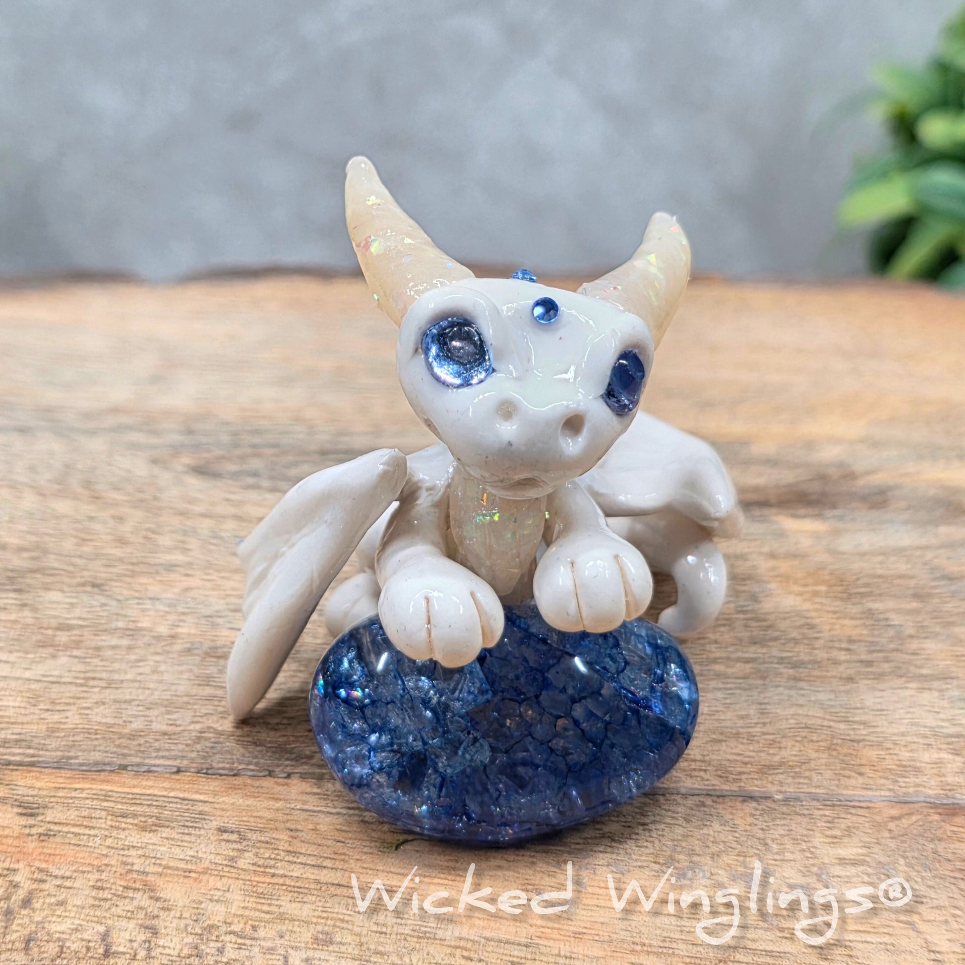A baby clay dragon sculpture with a crackled quartz against a wood background with greenery and text Wicked Winglings