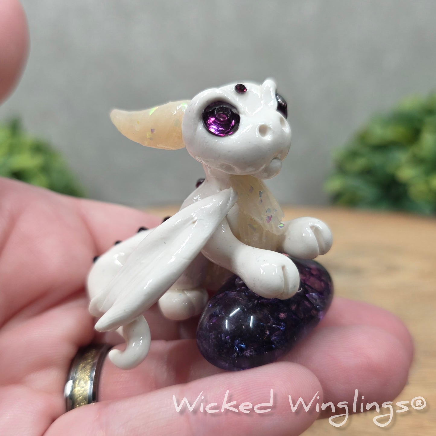 A hand holding a baby clay dragon sculpture with a crackled quartz against a wood background with greenery and text Wicked Winglings