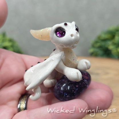 A hand holding a baby clay dragon sculpture with a crackled quartz against a wood background with greenery and text Wicked Winglings
