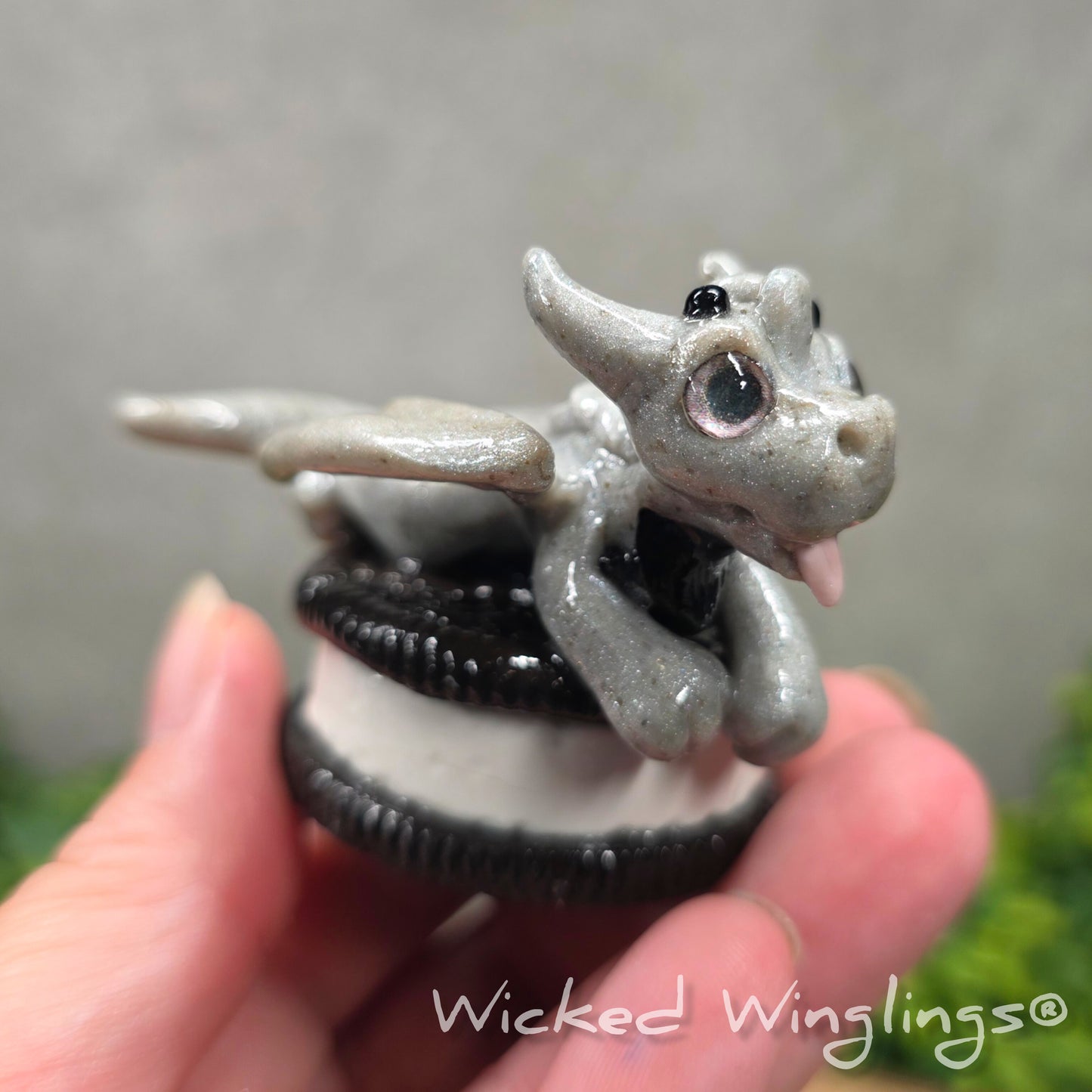 A hand holding a baby clay dragon with a cookie with a Wicked Winglings® watermark.