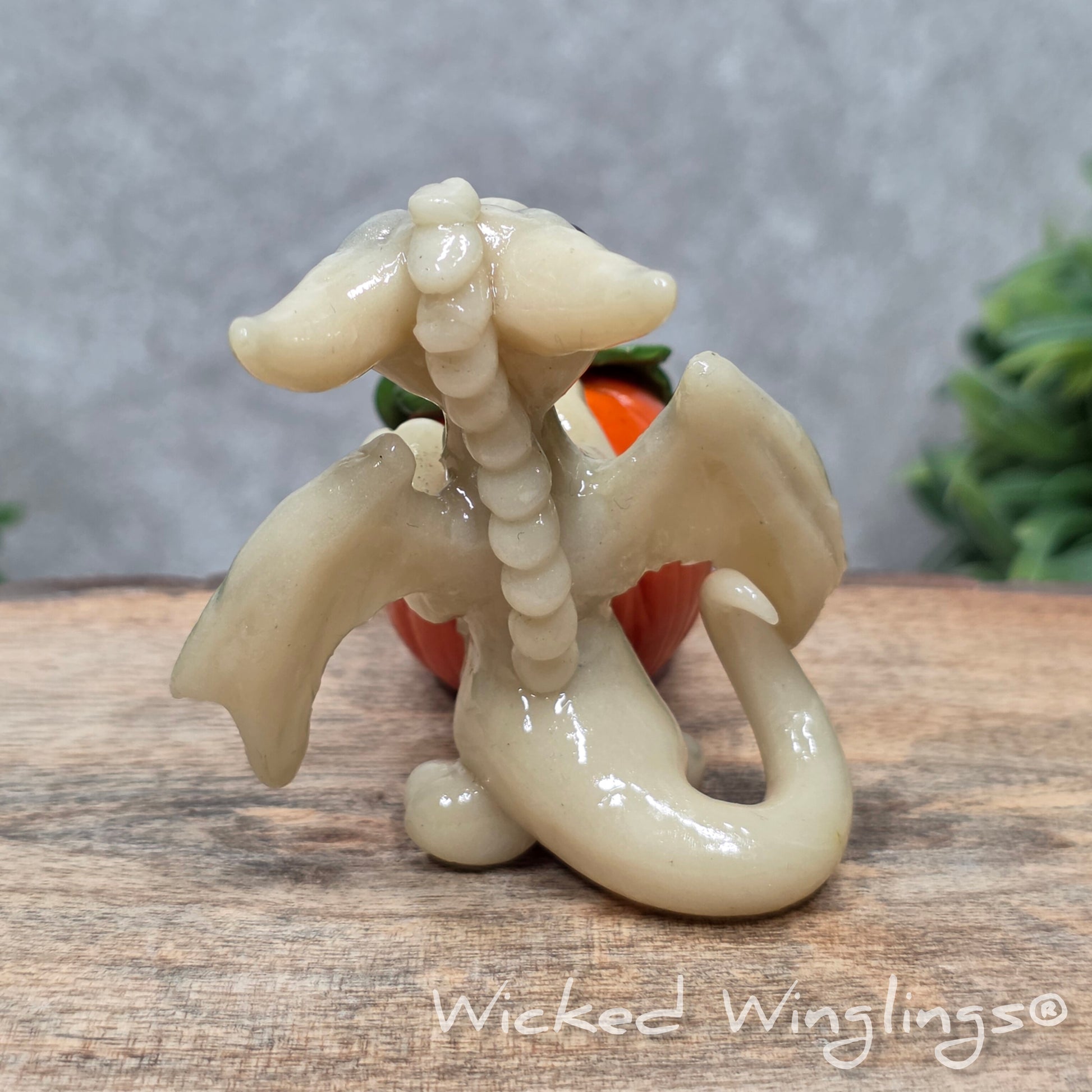 A baby clay dragon sculpture with a pumpkin against a wood background with greenery and text Wicked Winglings