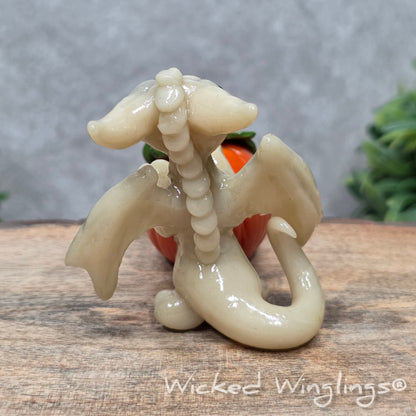 A baby clay dragon sculpture with a pumpkin against a wood background with greenery and text Wicked Winglings