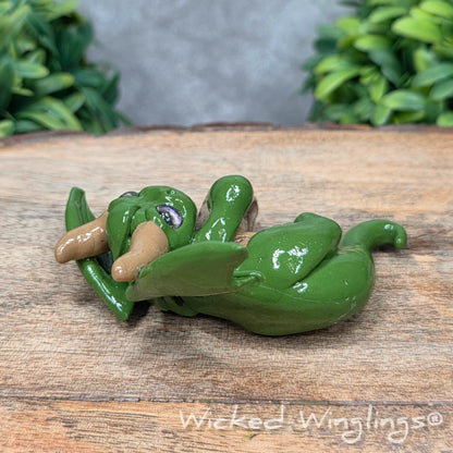A clay dragon with a skull on a wood surface with greenery. Includes text Wicked Winglings
