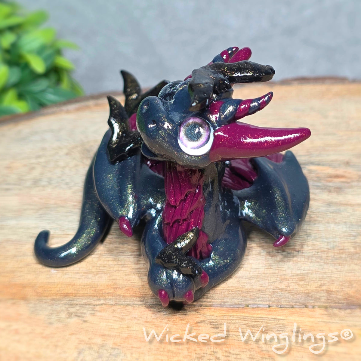 A clay dragon with bats sitting on a wood surface with greenery. Text reads Wicked Winglings.