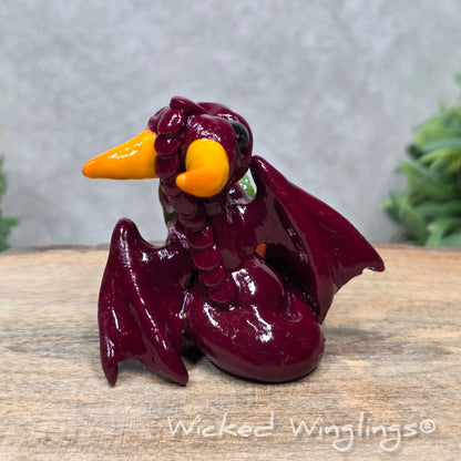 A baby clay dragon sculpture with a pumpkin against a wood background with greenery and text Wicked Winglings