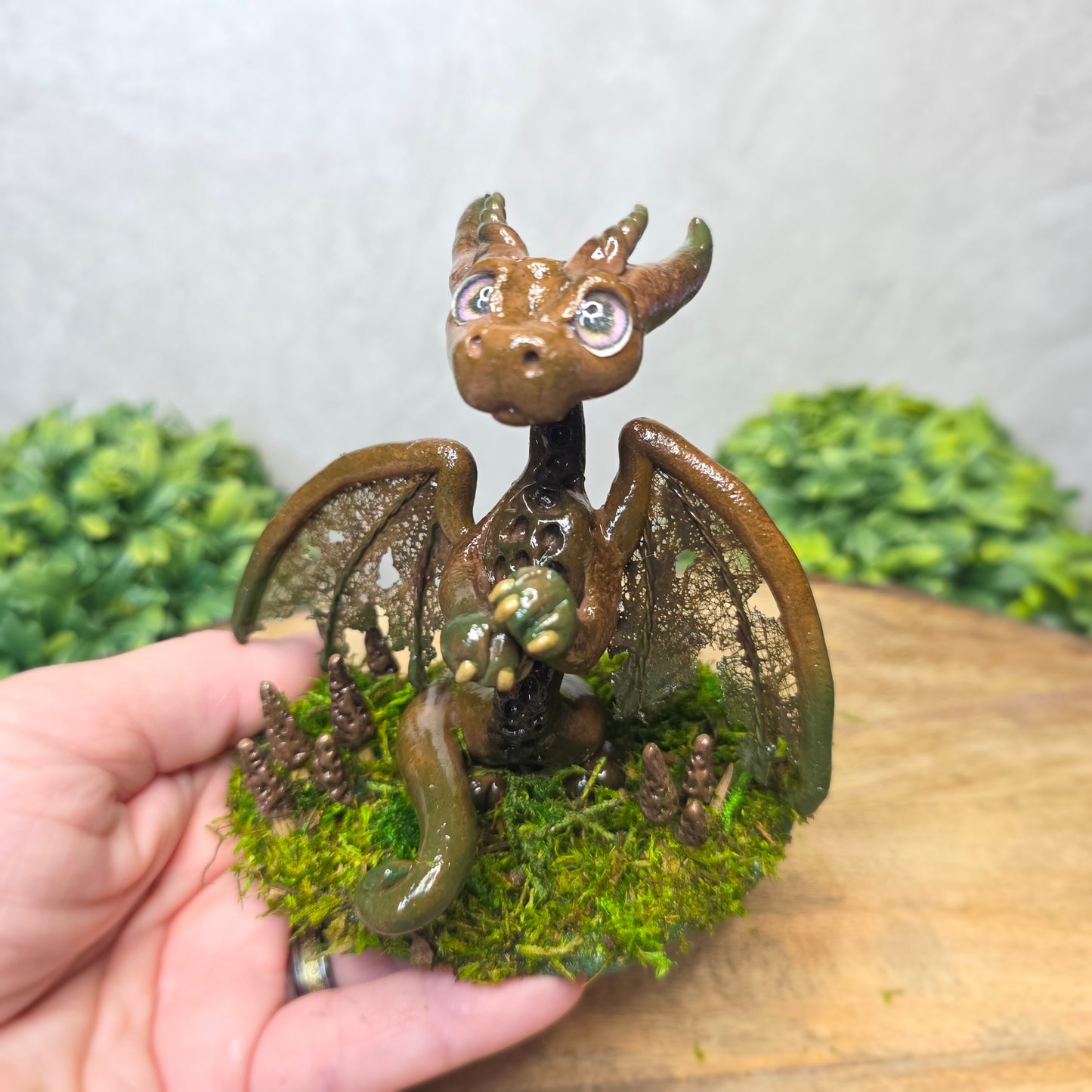 Hand holding a small dragon figurine with wings and a textured body against a blurred natural background.