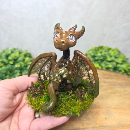 Hand holding a small dragon figurine with wings and a textured body against a blurred natural background.