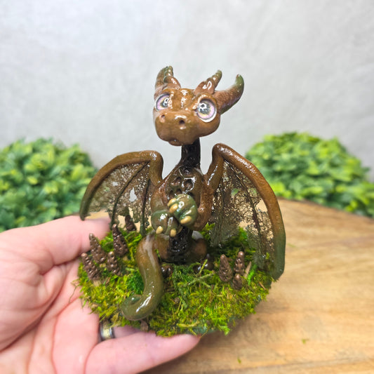 Hand holding a small dragon figurine with wings and a textured body against a blurred natural background.