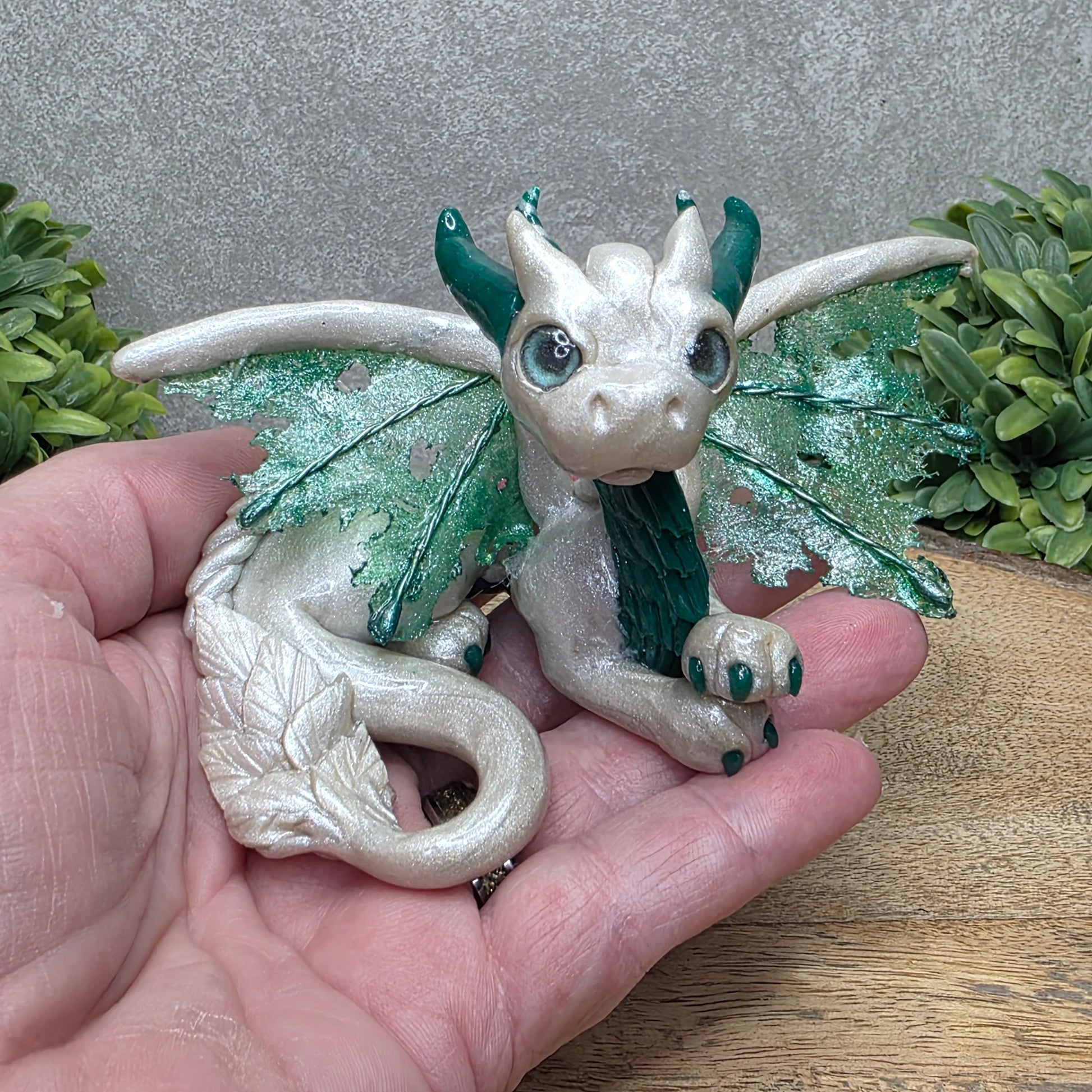 Hand holding a clay dragon against a wood surface with greenery.