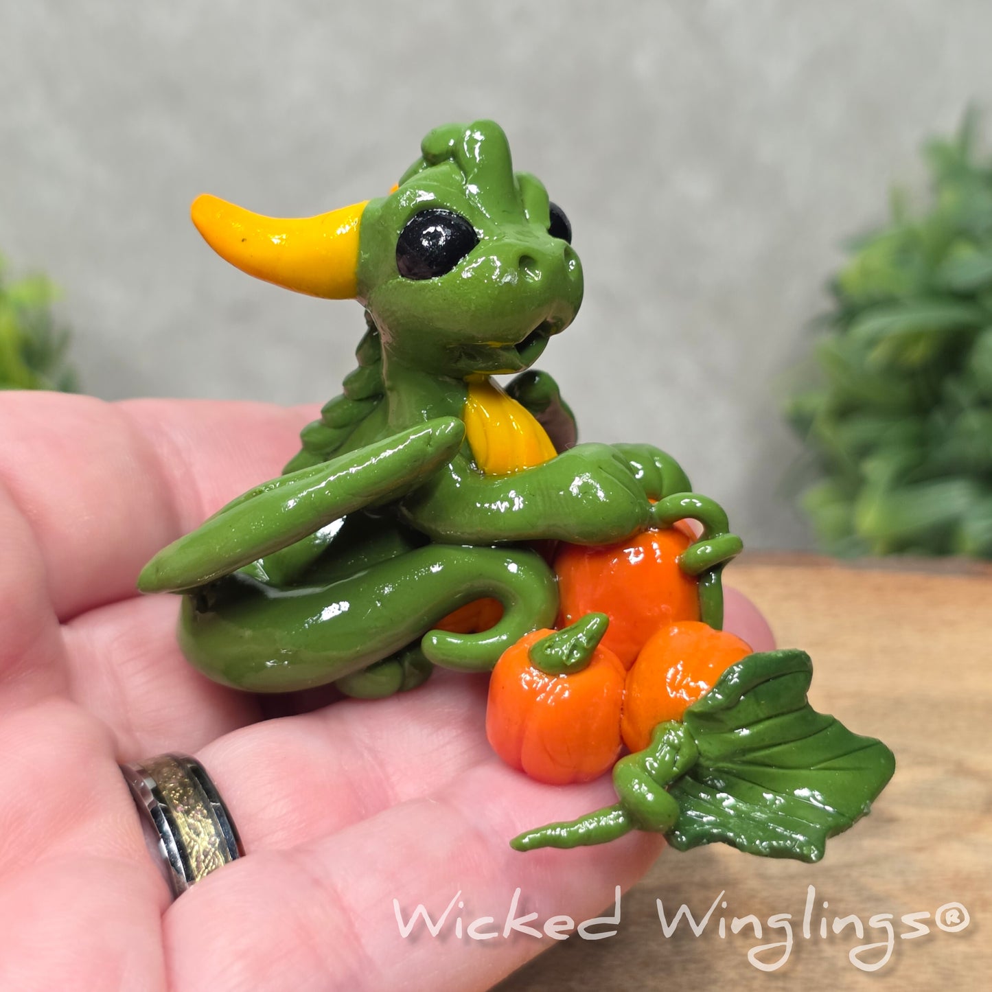A hand holding a baby clay dragon sculpture with pumpkins against a wood background with greenery and text Wicked Winglings