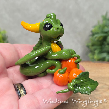 A hand holding a baby clay dragon sculpture with pumpkins against a wood background with greenery and text Wicked Winglings