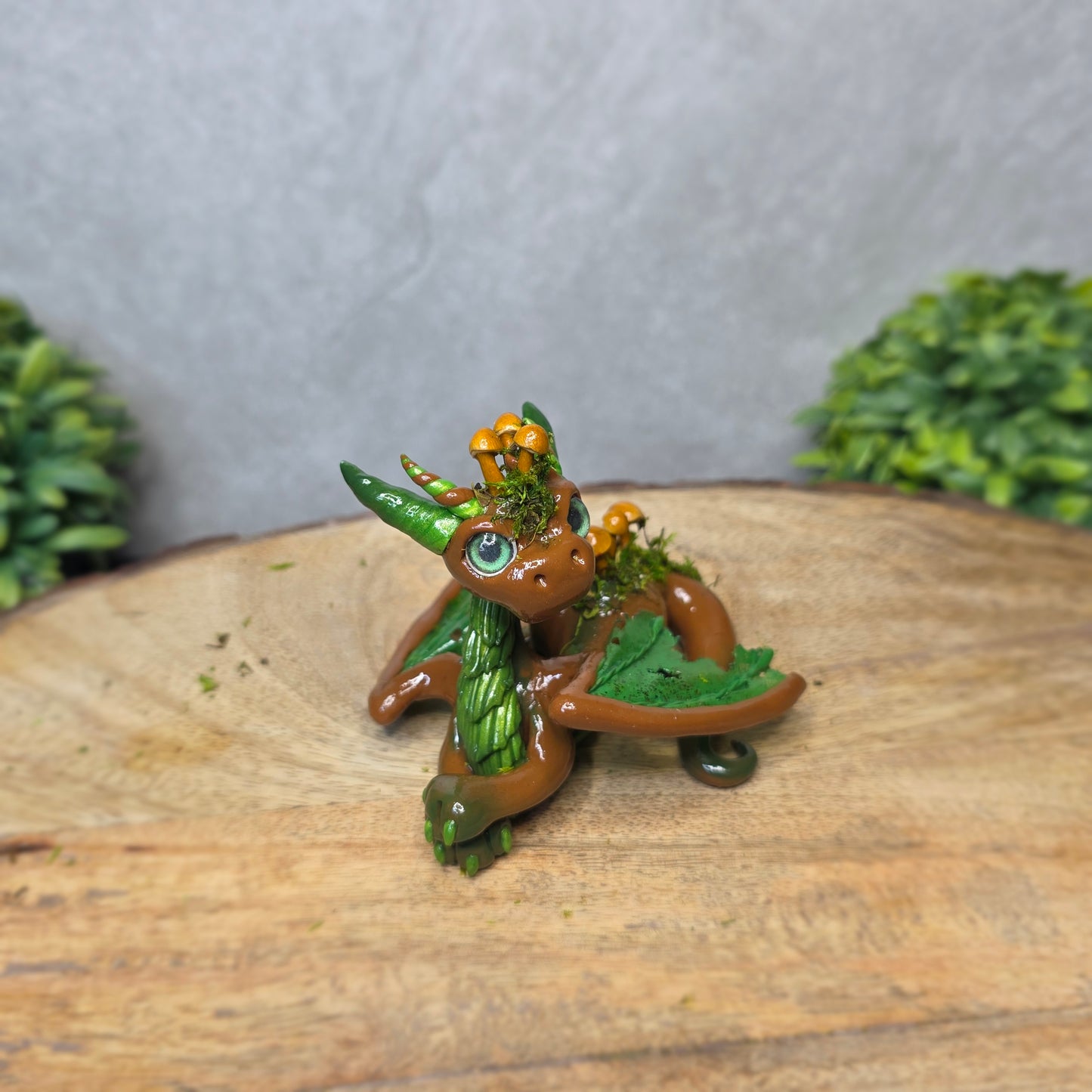 A clay dragon accented with moss and mushrooms against a wood surface with greenery.