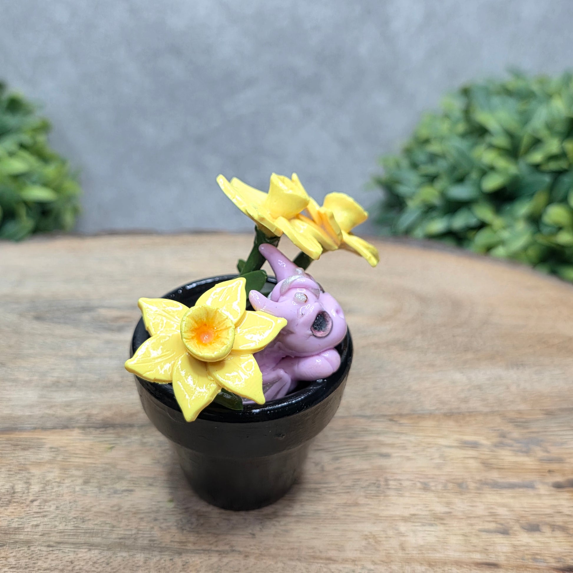 Side view of a clay baby dragon in a pot with daffodils against a wood surface with greenery.