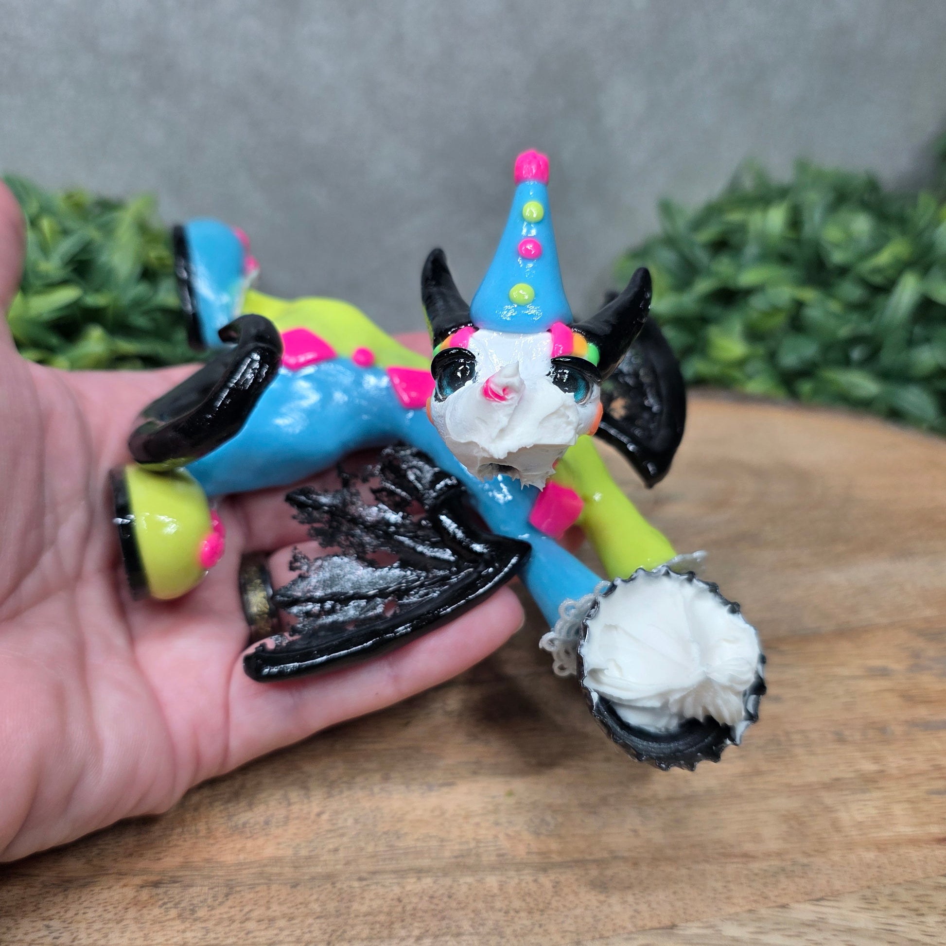 A hand holding a clown dragon with a cream pie on face against greenery
