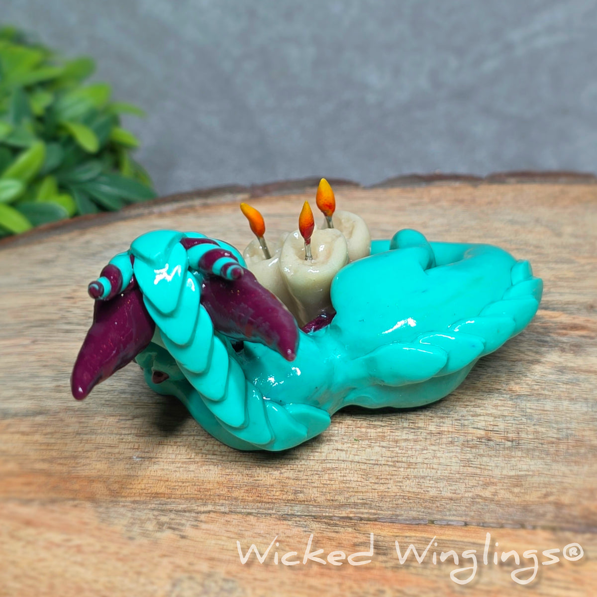 Back view of a clay dragon with candles against a wood surface with greenery. Text reads Wicked Winglings.