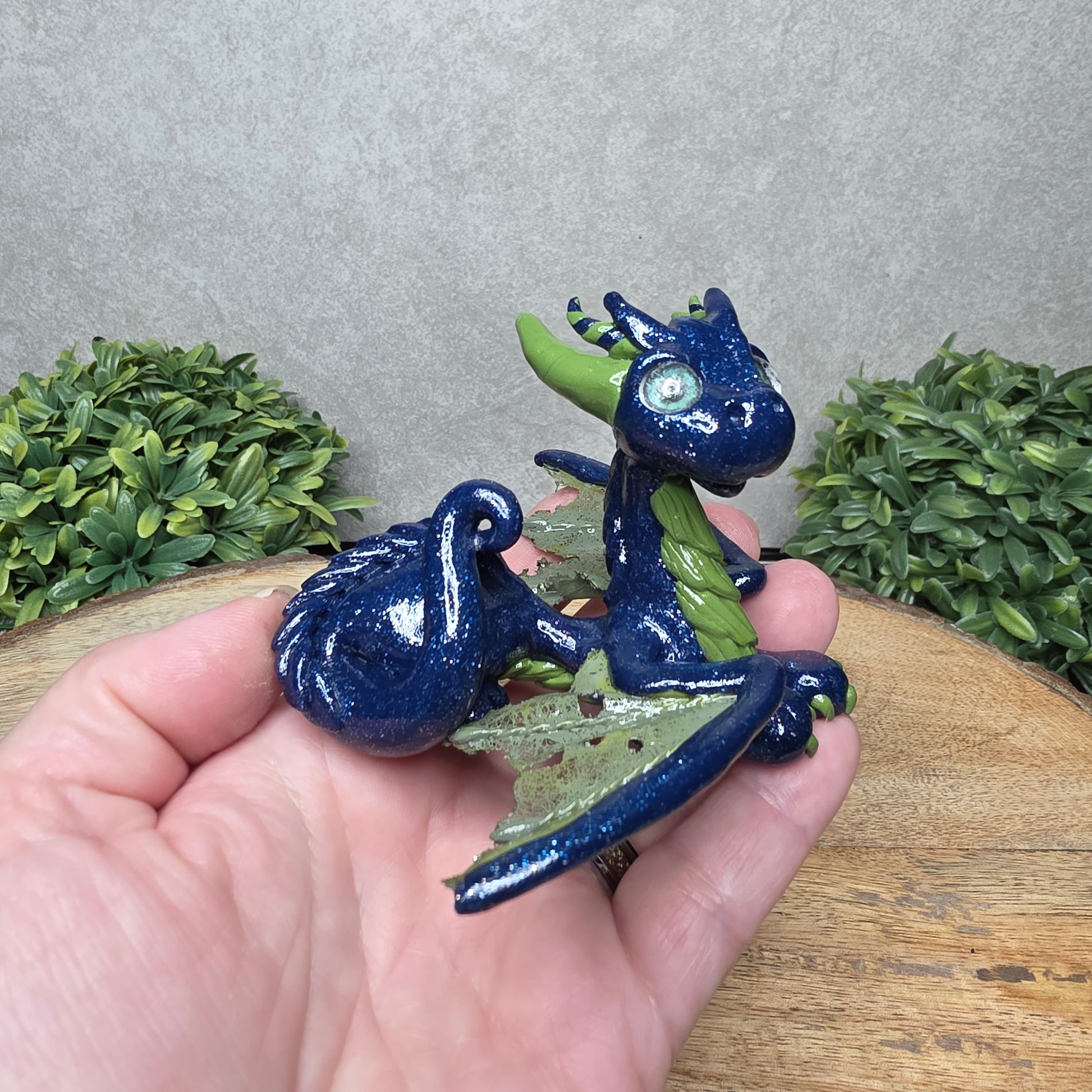 A hand holding a clay dragon against a wood background with greenery